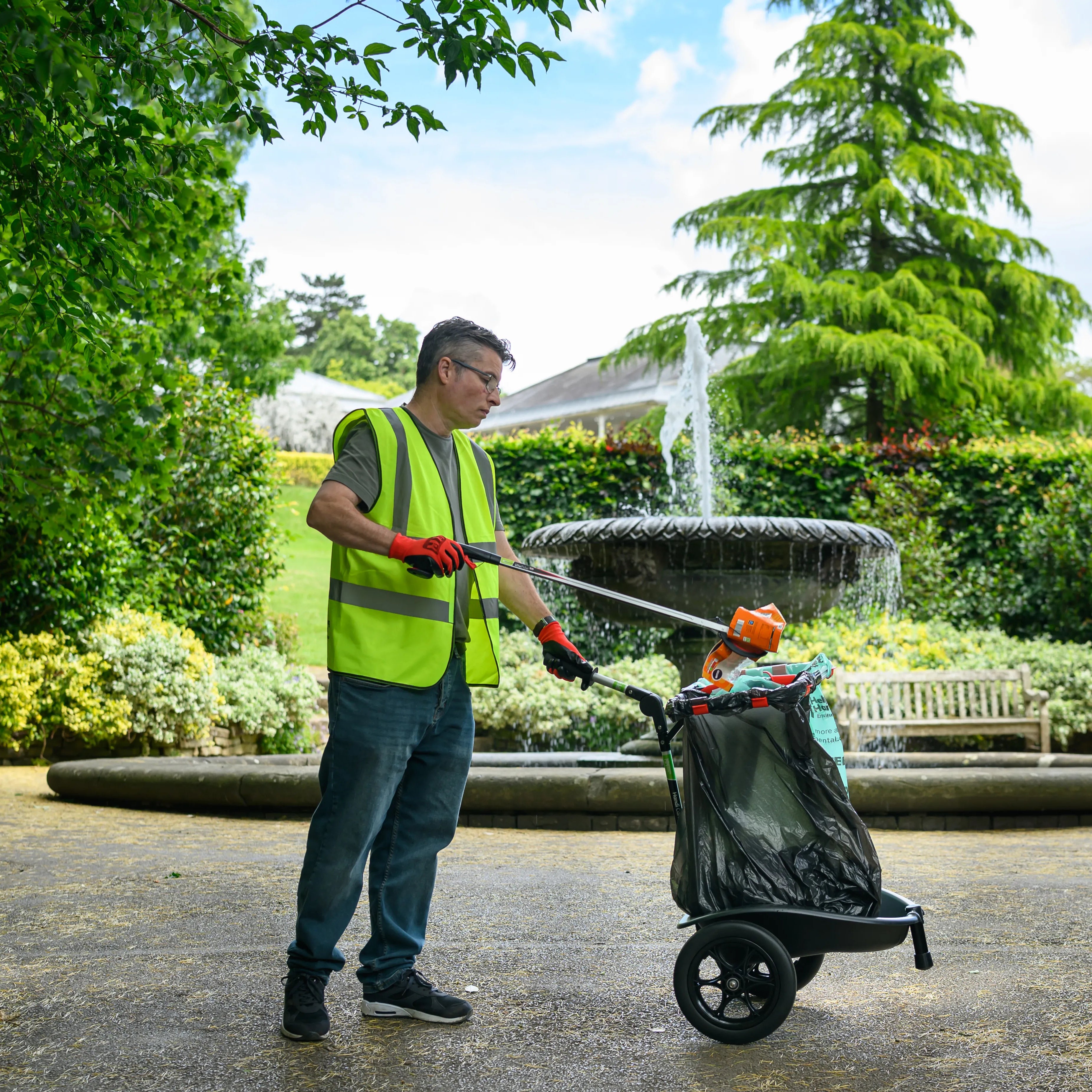 Person in a garden wearing a safety vest and pushing a litter cart.