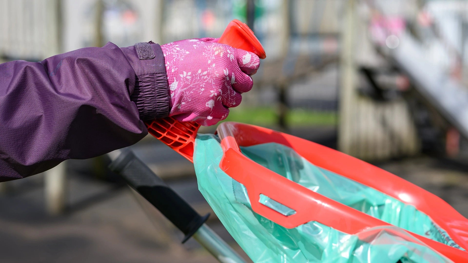 Person wearing purple jacket and pink gloves using a green recycling bin with red lid.