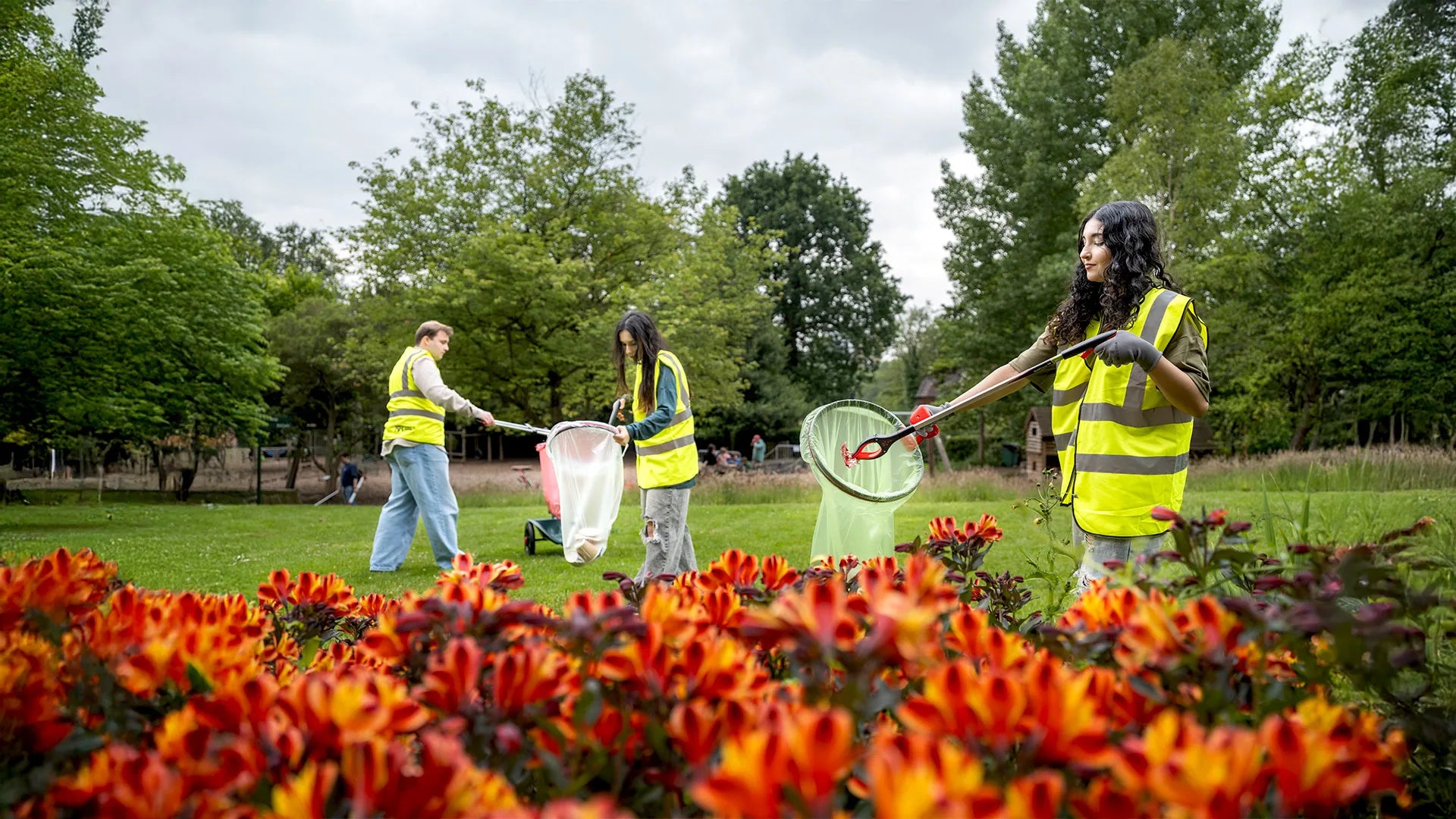 Three people in high-visibility vests cleaning up a park with litter hoops and litter pickers, surrounded by colorful flowers.