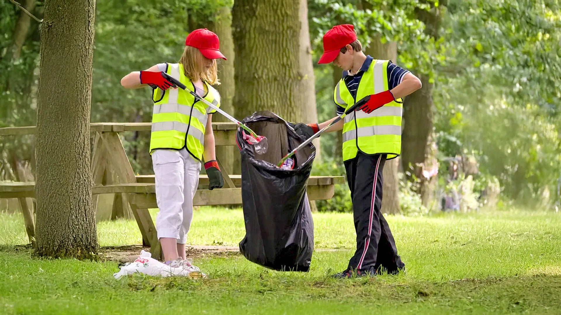 Two children in high-visibility vests picking up litter in a park.