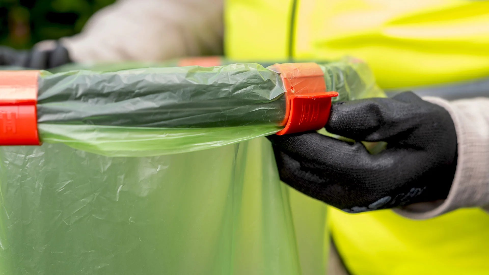 Person wearing a high-visibility vest and black gloves securing a green trash bag with a red clip.
