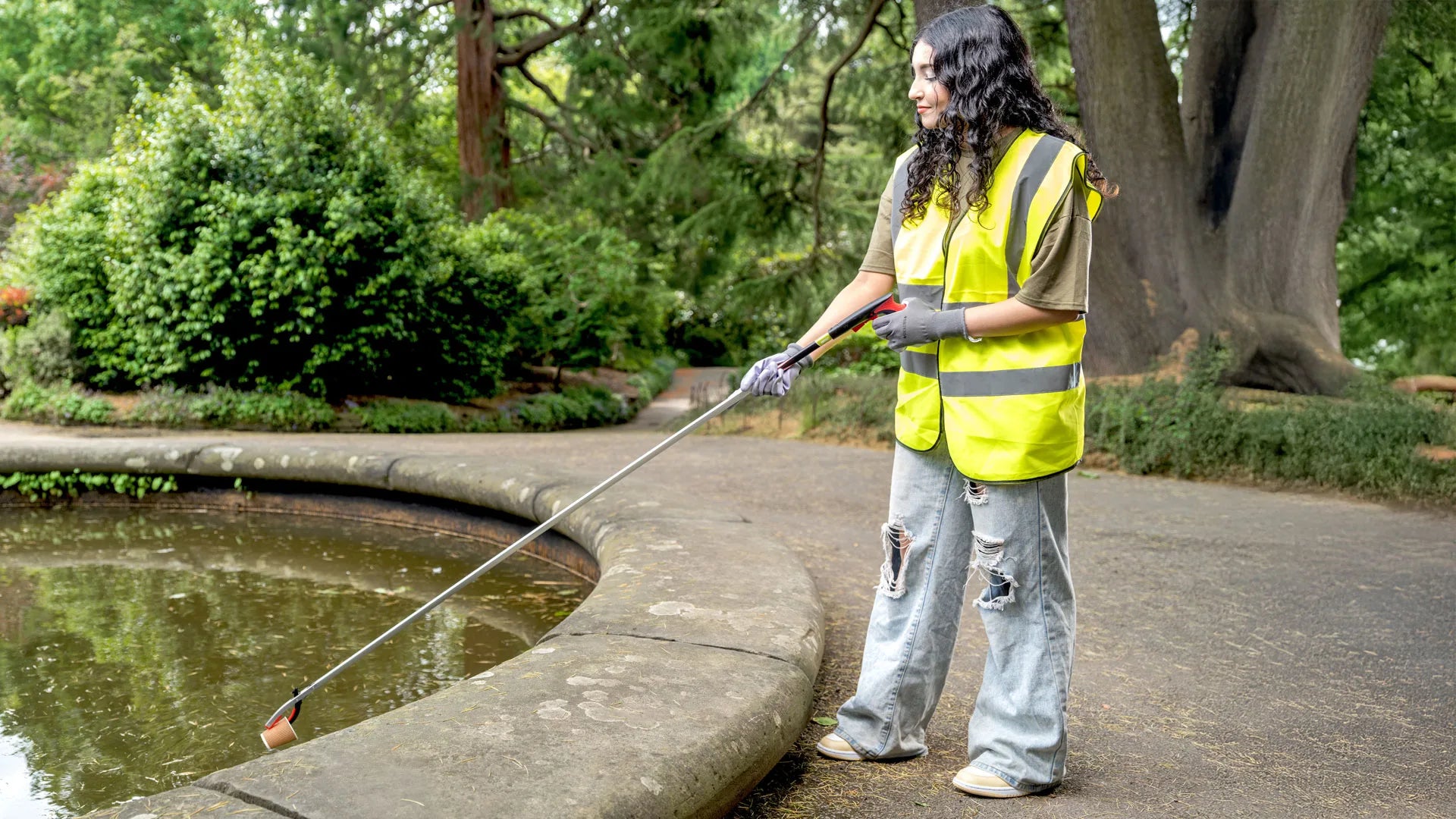 Person picking up trash from a pond with a long litter picker in a park setting.