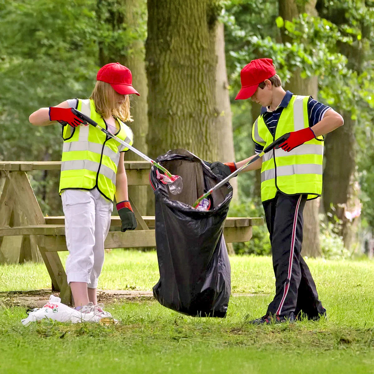 Two children in high-visibility vests picking up litter in a park.