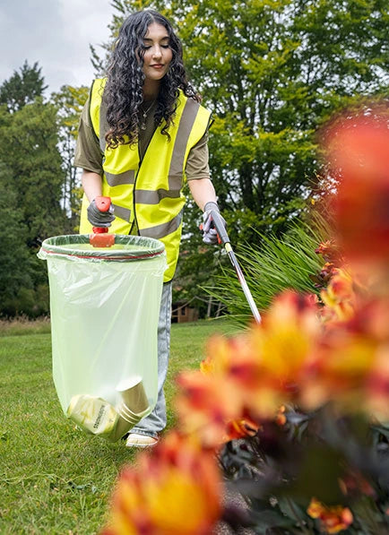 Volunteer in a yellow safety vest cleaning up trash in a park with flowers in the foreground.