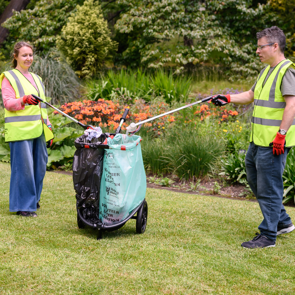 Two people in high-visibility vests using a trash picker to collect litter in a park.