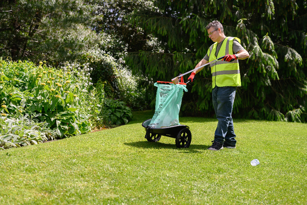 Person in a garden using a trash picker with a green bag on a grassy area.