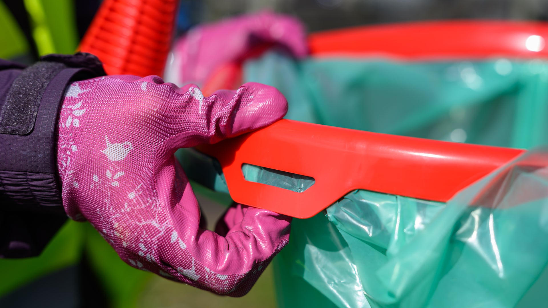 Hand wearing a pink gardening glove holding a red tool over a green trash bag.