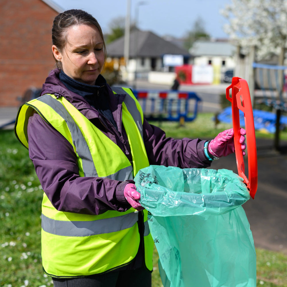 Person in a high-visibility vest holding a green trash bag and red tool outdoors.