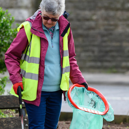 Person in a high-visibility vest holding a litter picker and green bag on a path.