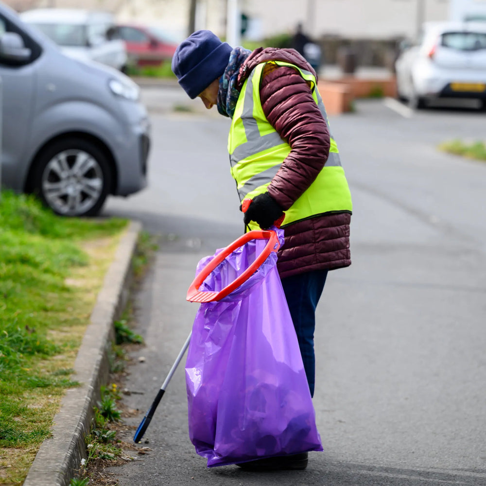 Person in high-visibility jacket and purple gloves cleaning up on a street.