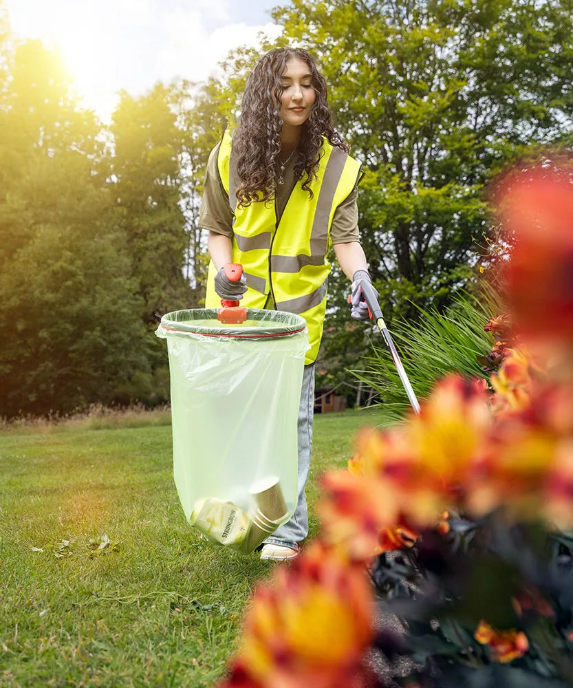 Person in a yellow safety vest cleaning up in a park with flowers in the foreground.