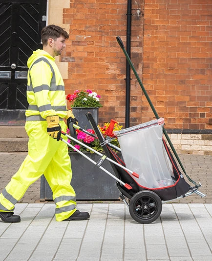Person in high-visibility clothing pushing a cleaning trolley with a trash bin on a sidewalk.
