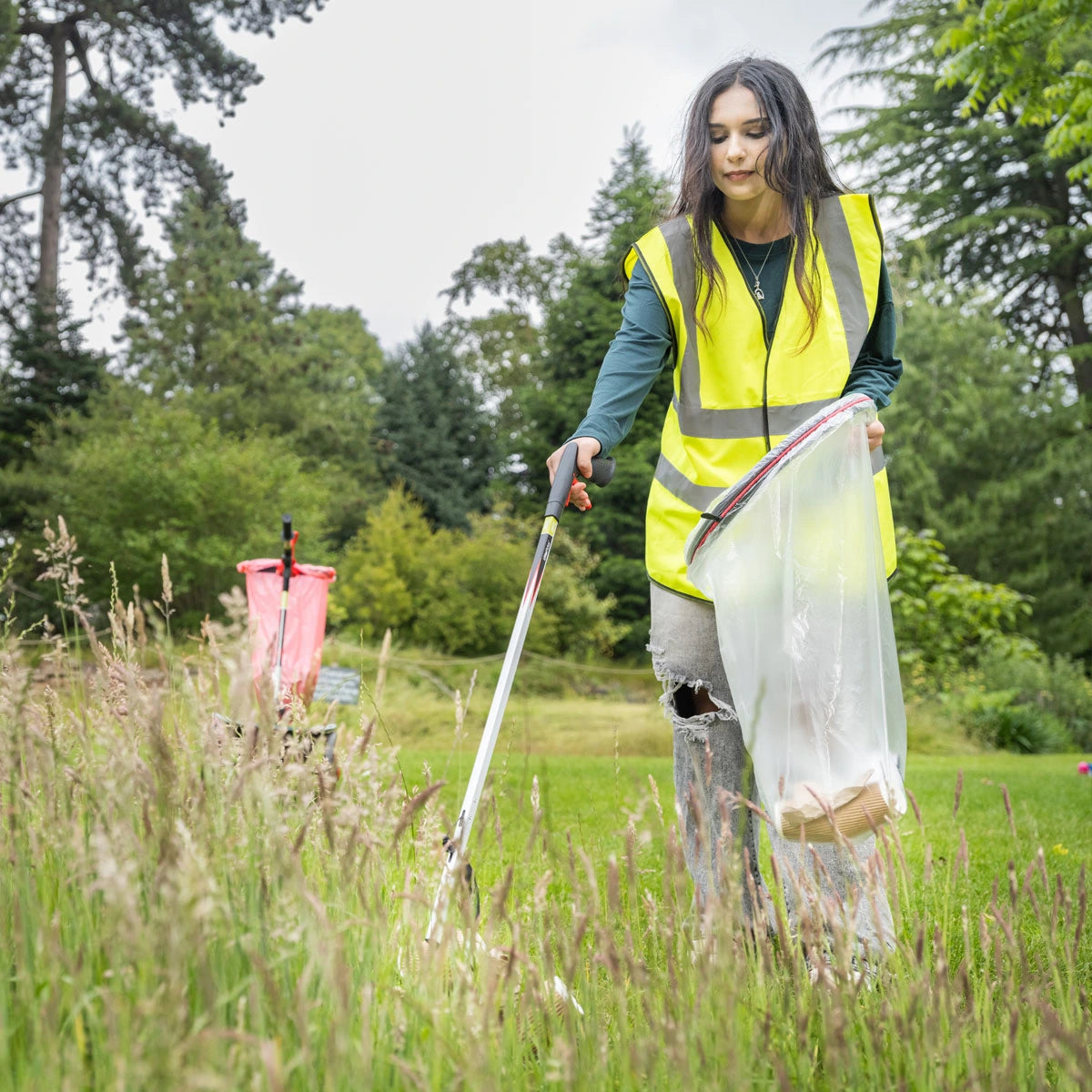Person in a yellow vest cleaning up in a grassy field with garden rubbish bag holder and litter picker.
