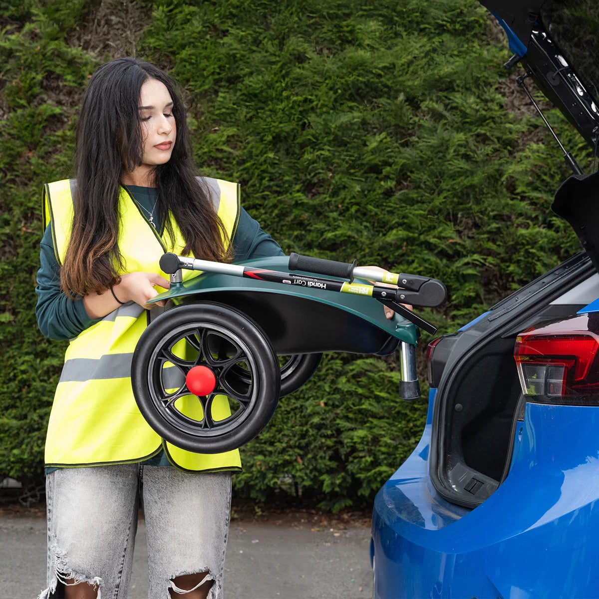 Person in a yellow safety vest loading Handi Cart Lite litter trolley into car trunk.