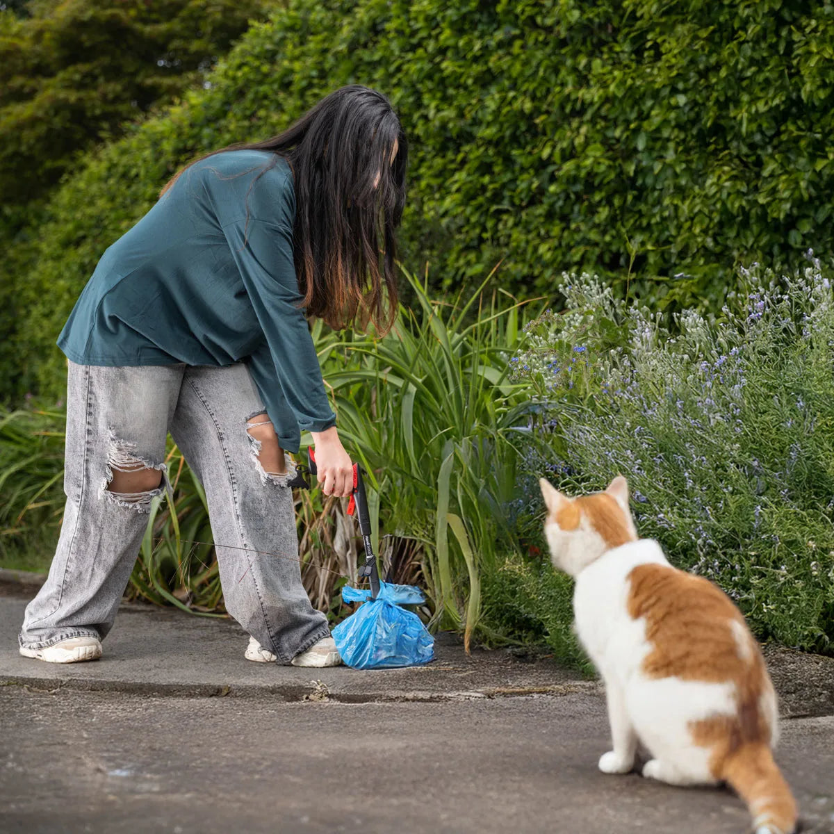 Person in a park picking up pet waste with the Handi Scoop Pro Pooper Scooper with a cat nearby.