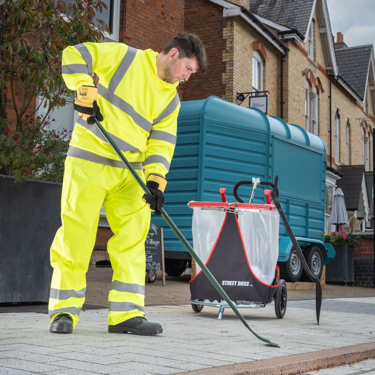 Person in high-visibility clothing using a street cleaning trolley on a residential street.