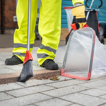 Person in high-visibility clothing cleaning a sidewalk with a broom and trash bag.
