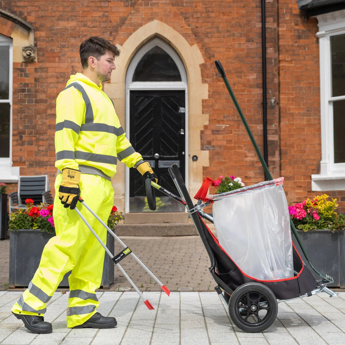 Person in high-visibility clothing pushing a cleaning cart with a bag on a pavement.